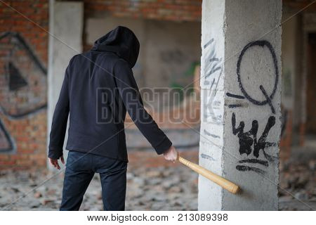 Street hooligan in black on the construction site hit a wooden bat with a gray column with graffiti.