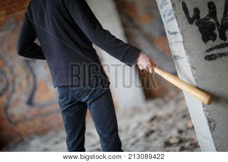 Street hooligan in black on the construction site hit a wooden bat with a gray column with graffiti.