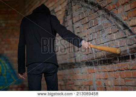 A street hooligan in a black hood at a construction site struck a brick red brick wall with a wooden bat.