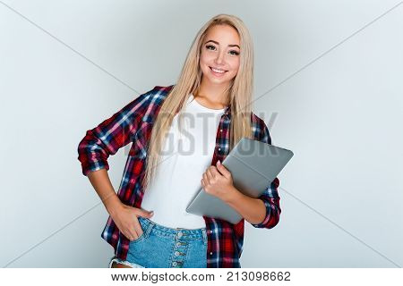 Candid student. Beautiful young woman with blond hair holding her laptop and looking at
camera with smile while standing against grey background