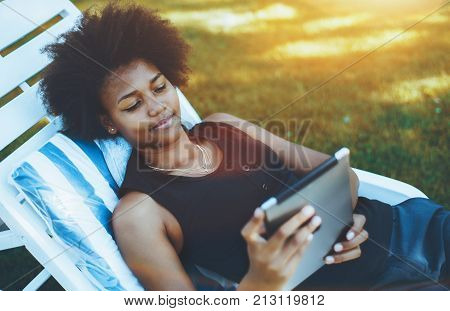 Close-up view of young charming Brazilian woman laying on deck chair and watching movie on her tablet pc; black cute curly girl on daybed in park chatting with her friend using digital tablet