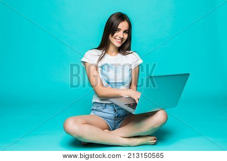 Happy Young Woman Sitting On The Floor With Crossed Legs And Using Laptop On Color Background