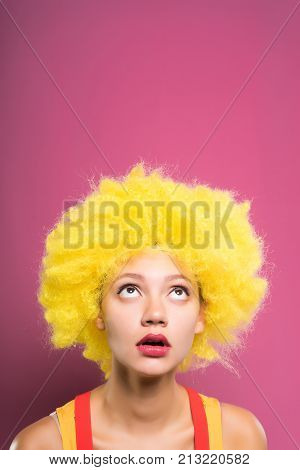 cute young girl in a yellow clownish wig looks up, on a pink background