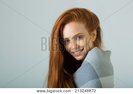 beautiful young girl with red hair warms up in a warm blanket on a light background