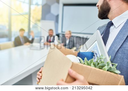 Unrecognizable man holding box of personal belongings looking back at several business people who fired him