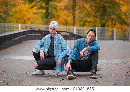 A young guy and a girl skateboarders, outdoors on a bright autumn day, the guy is sitting on a skateboard, the girl is sitting side by side.