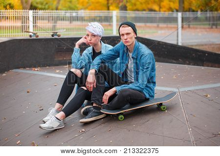 A young guy and a girl skateboarders, outdoors on a bright autumn day, sitting on a springboard of a man sitting on a skateboard, a girl next to her put her hand under his head.