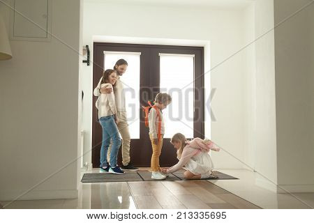 Little sister helping brother to tie shoes, girl fasten shoelaces of boy while parents looking at kids siblings son and daughter helping getting ready for walk together standing in hallway at home