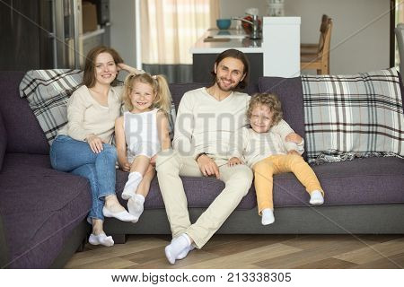 Happy smiling family of four sitting on sofa looking at camera, young couple with kids boy and girl relaxing on couch at home, parents with children posing in living room, mortgage loan, insurance