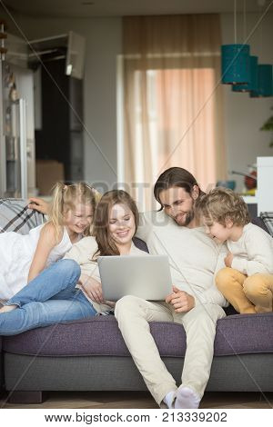 Family with children having fun using laptop sitting on sofa at home, parents and son daughter relaxing on couch holding computer, man woman with kids smiling watching funny video online, vertical