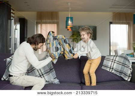 Pillow fight between father and little son in living room, happy dad and child boy playing on sofa fighting with cushions, daddy laughing having fun with his kid together, leisure activity at home
