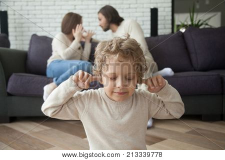 Little boy puts fingers in ears not to hear parents fighting at background, upset tired son suffering from mom and dad arguing, parental conflicts hurt kid, family divorce effect on children concept