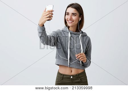 Portrait of good-looking cheerful young caucasian girl with dark long hair in casual gray hoodie and jeans smiling brightly, holding smartphone in front of face to make selfie during listening to fave song