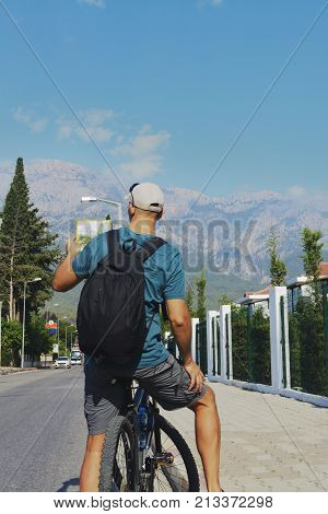 a man stands on the road in the village and looks at the tourist map dressed in a T-shirt and cap. a man with a backpack stands on a background of high mountains.