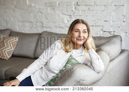 Indoor portrait of attractive happy 60 year old senior woman with pleasant smile relaxing on grey sofa in her modern apartment looking in anticipation while waiting for her children or grandchildren
