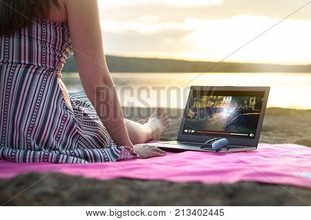 Young woman streaming a movie with laptop computer on beach at sunset. Watching film stream on imaginary online service outdoors. Video player on screen.