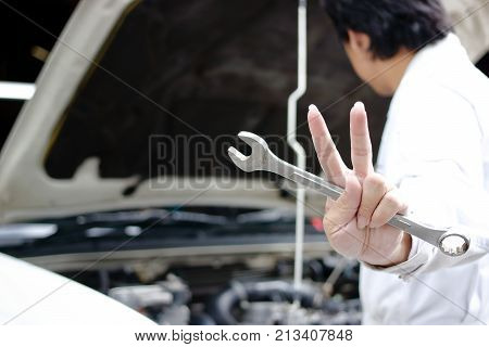 Hands of confident young mechanic holding wrench and raising two fingers up it is shows fight with job against car in open hood at the repair garage.