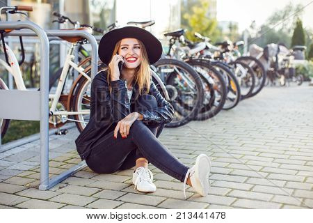Stylish hipster girl wears black leather jacket sitting on ground and holding phone before bike stop