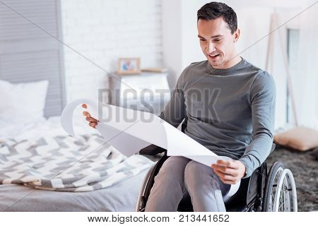Analyzing sketch. Cheerful disabled keeping smile on his face and bowing head while looking at draw