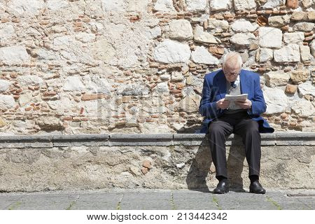 TAORMINA, ITALY. April 3, 2015: Old man sitting reading a newspaper. Ancient wall of a church in the center of Taormina in Sicily, Italy.