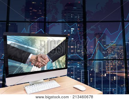 the computer on the wood table with Hand shake between businessman at the screen on the trading graph over the blurred photo of cityscape backgroundElements of this image furnished by NASA