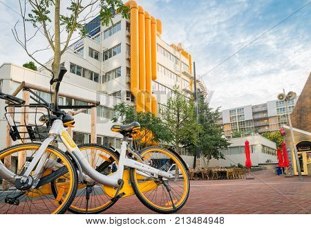 ROTTERDAM HOLLAND- AUGUST 23 2017; Two yellow and white share bikes leaning against tree in front of city library building with bright yellow external air-conditioning ducts in city square