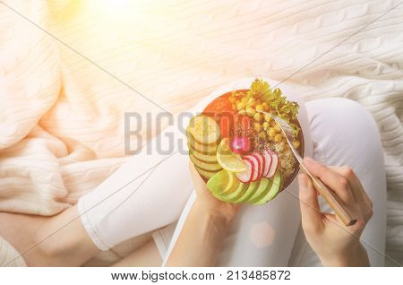 Girl in white jeans holds in hands fork, vegan breakfast meal in bowl with avocado, quinoa, cucumber, radish, salad, lemon, cherry tomatoes, chickpea, chia seeds. Top view, copy space. Clean eating.