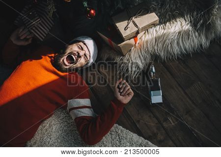 Another picture of an adult guy lying on the floor near pine tree with tree toys and Christmas gifts this sunny morning. After parting all night long this man has a terrible hangover. And he is not happy the sunlight makes him get up. Cut view
