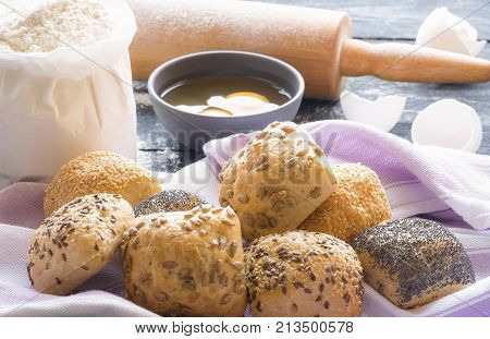 Buns with seeds and ingredients - Food theme image with homemade bread rolls with sesame poppy seeds sunflower and flax seeds on a towel and the ingredients for their baking in the background.