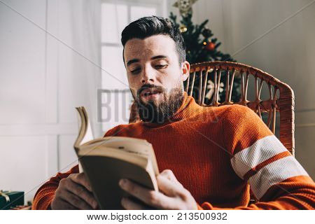 Close up of attractive young man sitting in the rocking chair and reading a book. He is sitting besides the green Christmas pine, full of tree Christmas toys. It's so nice to do some cozy things almost at Christmas eve. Cut view