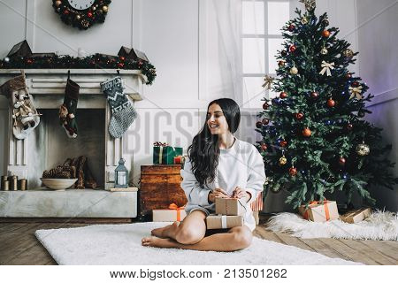 Beautiful girl in white sweater is sitting on big cozy carpet and holding boxes of gifts for her beloved. On the back there are a white fireplace covered with garlands and socks and green pine tree full of Christmas toys. Christmas is coming.