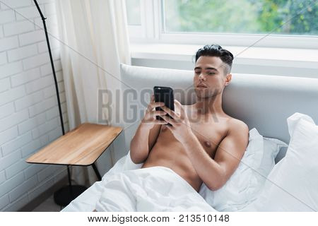 The guy lies in a white bed and looks something on the screen of his black smartphone. Behind him is a window overlooking the street. He lies on the bed alone.