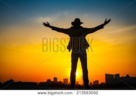 Traveler silhouette with a backpack and cowboy hat stand on hill top. City in the background. Sun sets over horizon. Man looks ahead, raise his arms up. Travel, holidays, advanture, success concept.