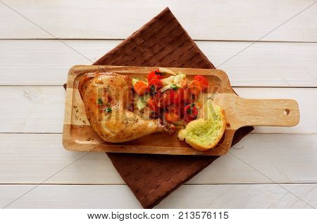 Chicken leg roasted with fried vegetables and garlic bread in wood board with brown napkin on white table