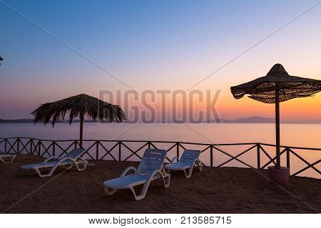 Beach umbrellas and sun loungers on the sand beach on the background of calm water surface . Red Sea at sunrise. Sharm el-Sheikh. Egypt. Concept of tourism and beach recreation