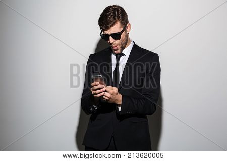 Close-up photo of amazed bearded man in black suit holding mobile phone, isolated on white background
