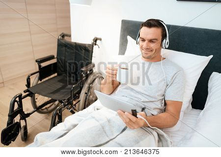 The invalid lies in bed and looks something on the tablet. He smiles. because he is happy. Next to his bed is his black wheelchair. It lies on a large white step.