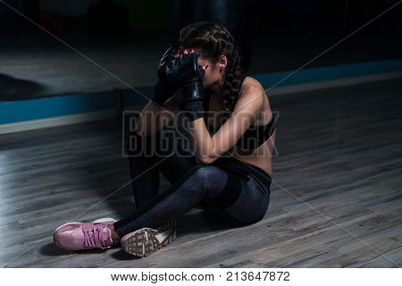 Upset young fighter boxer girl wearing boxing gloves siting on the floor after loosing fight. She covered her face with hands.