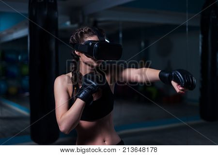 Young fighter boxer fit girl in VR glasses wearing boxing gloves in training. Boxing game in virtual reality 360 degrees. Futuristic gaming in real gym