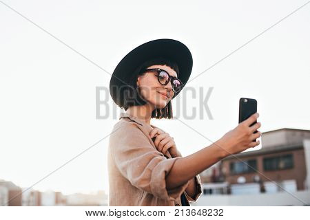 Cute and pretty fashion trendy and hipster millennial woman or girl makes selfie on smartphone camera to share on internet social media channels self absorbed new generation of young people
