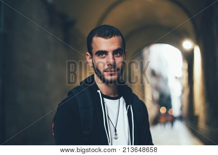 Handsome attractive young man in 20s student or professional freelancer looks into camera stands in dark street or alley in Barcelona exchange education program confident and calm natural male beauty