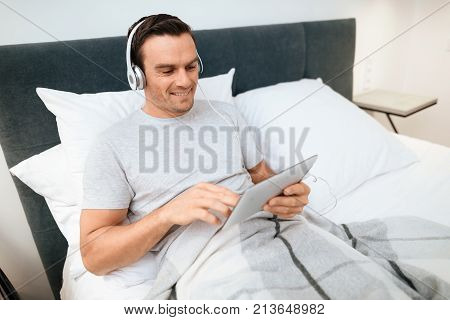 The invalid lies in bed and looks something on the tablet. He smiles. because he is happy. Next to his bed is his black wheelchair. It lies on a large white step.