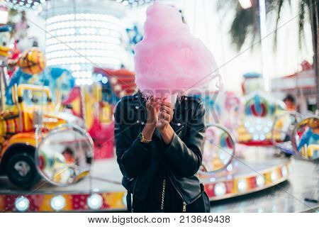 Happy attractive and beautiful young woman or female teenager with blonde hair wears black leather jacket stands in front of attraction ride at festival or carnival and holds huge pink cotton candy