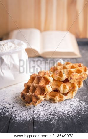 Homemade belgian waffles - Culinary theme image with a bunch of delicious freshly baked belgian waffles surrounded by wheat flour and an open cookbook on a wooden table.