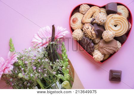 Heart shaped box with cookies and flowers - Gifting theme image with a red heart shaped box full of delicious sweets and a bouquet of flowers on a pink background. A greeting card idea.