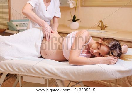 the massage therapist makes a belly massage to a pregnant young woman