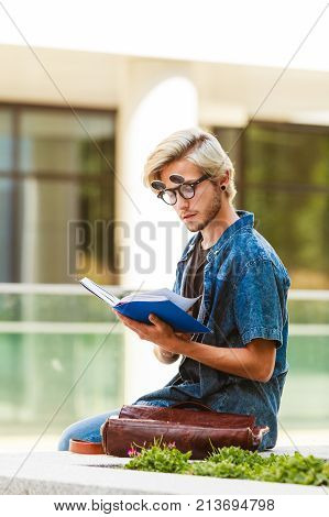 Male fashion student concept. Guy holding and studying from notebook wearing jeans outfit and eccentric sunglasses sitting on white ledge next to modern building