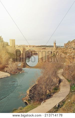 An Arch Alcantara Bridge Over The Tagus River And Its Reflection In Water And A Path Along The River