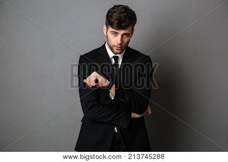 Serious hansome guy in black suit checking time on his wristwatch, looking at camera, isolated on gray background