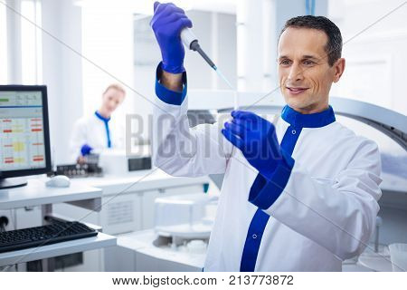 One more drop. Skillful attractive male madical worker dropping medication with a pipette into vial which holding in his hand  while standing next to the computer